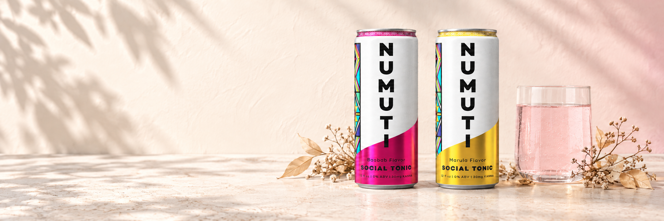 NuMuti Baobab and Marula cans beside a glass of sparkling pink tonic on a sunlit stone surface.